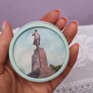 May include: A round, light blue plastic trinket box with a picture of a statue on top of a tall, rectangular base. The statue is of a person in a dark suit standing with their arm raised. The background is a light blue sky with some clouds.
