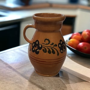 Puede incluir: Una jarra de cerámica marrón con asa, decorada con diseños florales negros y rayas rojas. La jarra está sobre una superficie blanca. El fondo está borroso, pero parece ser una cocina.