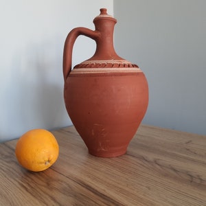 Op de afbeelding: Een terracotta kan met een wit ontwerp en een ronde sinaasappel op een houten tafel.
