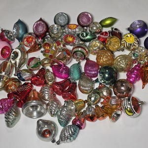 Könnte beinhalten: Eine Sammlung von Vintage-Weihnachtsschmuck in verschiedenen Formen und Farben, darunter Rosa, Silber, Rot und Gold. Die Ornamente bestehen aus Glas und haben verschiedene Designs, wie Streifen, Wirbel und Glocken. Eine festliche Auswahl für die Weihnachtsdekoration.