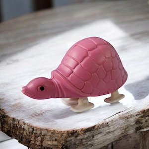 Op de afbeelding: Een roze wind-up speelgoed schildpad met een gedetailleerde schild en witte wielen. De schildpad staat op een houten oppervlak.