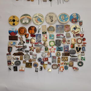 May include: A collection of vintage enamel pins featuring a variety of designs, including animals, sports, and political figures. The pins are arranged in rows on a white surface. Some of the pins have Cyrillic text, indicating they are from the Soviet Union.