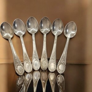 May include: Six silver teaspoons with ornate designs on the handles. The spoons are arranged in a row on a reflective surface.