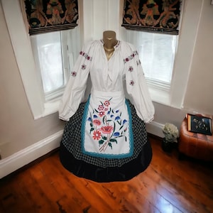 Op de afbeelding: Traditionele witte blouse en rok ensemble. De blouse heeft geborduurde bloemmotieven in rood en paars. De rok heeft een ruitpatroon met een zwarte onderkant. Een schort met bloemborduurwerk in rood, blauw en groen is ook inbegrepen.