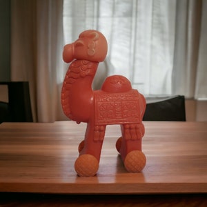 May include: A pink plastic camel toy with a decorative saddle and wheels. The camel has a detailed head with a patterned design.