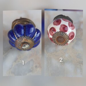 Puede incluir: Dos pomos de armario de cerámica. El primer pomo es azul con un borde dorado y el segundo pomo es blanco con lunares rojos y un borde dorado.