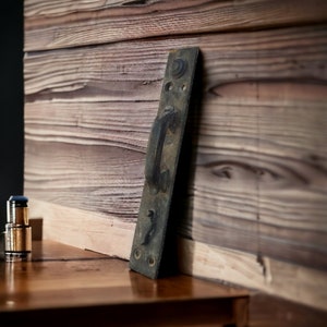 Puede incluir: Una manija de puerta de metal oxidado vintage con un acabado negro y un diseño simple. La manija está unida a una puerta de madera.