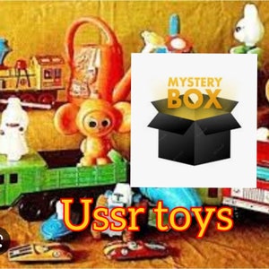 Puede incluir: Una colección de juguetes soviéticos vintage, que incluyen un tren de juguete, una regadera, un juguete de peluche y un coche de juguete. Los juguetes están dispuestos en una mesa con un cartel que dice "Mystery Box" y "USSR toys".
