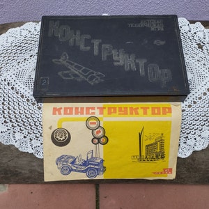 Op de afbeelding: Een vintage zwarte doos met de tekst "Конструктор" in witte letters. De doos is open en toont een gele instructiepagina met een zwart-witte illustratie van een speelgoedauto en de tekst "Конструктор" in rode letters.