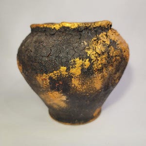 Puede incluir: Una pequeña maceta de cerámica negra con detalles dorados. La maceta tiene una superficie rugosa y texturizada y está ligeramente agrietada.