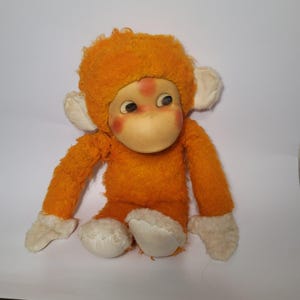 Puede incluir: Juguete de mono de peluche naranja con cara blanca y ojos grandes. El mono está sentado con los brazos extendidos.