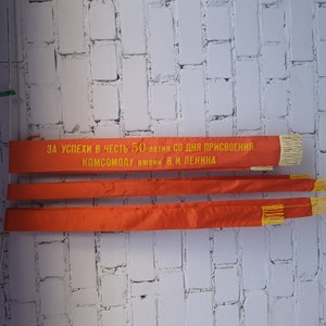 May include: Three red fabric banners with yellow text that reads "ЗА УСПЕХИ В ЧЕСТЬ 50-летия СО ДНЯ ПРИСВОЕНИЯ КОМСОМОЛУ имени В. И. ЛЕНИНА".