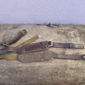 May include: A brown leather strap with a metal buckle and a brown canvas strap. The straps are lying on a wooden surface.