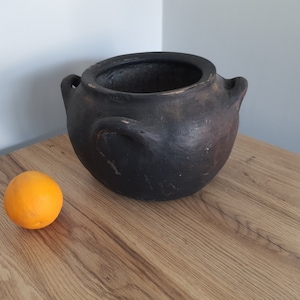 Può includere: Un vaso di terracotta nero con due manici su una superficie di legno accanto a un'arancia.