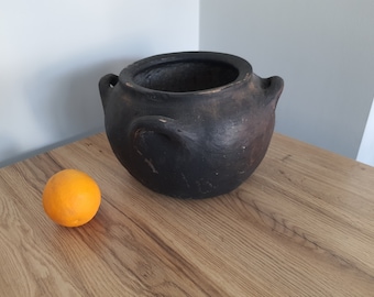 May include: A black clay pot with two handles sits on a wooden surface next to an orange.