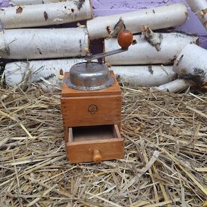 May include: A vintage wooden coffee grinder with a metal grinding mechanism and a drawer. The grinder has a crank handle and a wooden knob on the drawer. The grinder is sitting on a bed of straw.