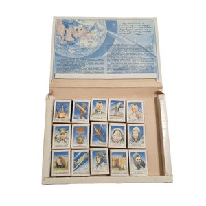 Puede incluir: Un juego de cajas de cerillas vintage con 16 cajas de cerillas individuales que muestran imágenes de exploración espacial, astronautas y naves espaciales. Las cajas de cerillas están dispuestas en una cuadrícula dentro de una caja de cartón más grande. La caja tiene un fondo azul y blanco con texto en ruso.
