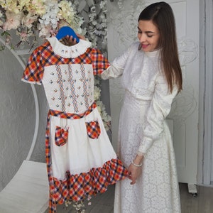 May include: A white dress with a lace pattern and long sleeves. The dress is worn by a person standing next to a white dress with a red, orange, and black plaid pattern. The plaid dress has a white collar and embroidered details.