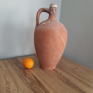 Puede incluir: Una jarra de barro cocido con una boca ancha y un asa. La jarra está sentada en una mesa de madera junto a una pequeña naranja.