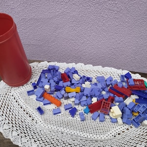 May include: A large pile of colorful plastic building blocks in various shapes and sizes. The blocks are mostly purple, blue, red, and yellow. The blocks are sitting on a white crocheted doily.