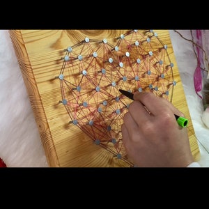 Könnte beinhalten: Ein Holzbrett mit einer Herzform, die mit Nägeln und roter Schnur erstellt wurde. Eine Hand hält einen grün-schwarzen Stift und fädelt die rote Schnur zwischen die Nägel.