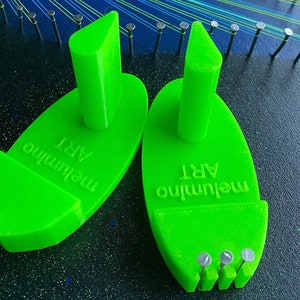 Könnte beinhalten: Zwei hellgrüne String-Art-Werkzeuge mit dem Text "melumino ART" sind abgebildet. Jedes Werkzeug hat einen zentralen Pfosten und eine gebogene Basis mit kleinen Nägeln. Die Werkzeuge liegen auf einer dunklen Oberfläche mit String-Art in Arbeit.