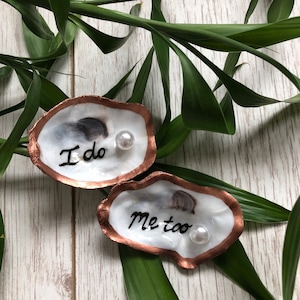 May include: Two oyster shells with copper-coloured edges and white interiors. One shell has the words "I do" and the other says "Me too" in black script. Each shell has a small pearl. Green leaves are in the background.