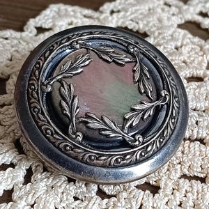 Könnte beinhalten: Ein runder, antiker silberner Kompaktpuder mit einem verzierten Blattdesign am Rand. Die Mitte zeigt eine perlmuttartige, irisierende Oberfläche. Der Kompaktpuder liegt auf einer cremefarbenen, gehäkelten Tischdecke.