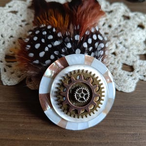 Puede incluir: Broche de estilo steampunk con un diseño en capas. El broche circular tiene un anillo exterior de nácar y marrón, un engranaje de latón en el centro y un pequeño engranaje en el medio. Plumas moteadas marrones y blancas detrás del broche.