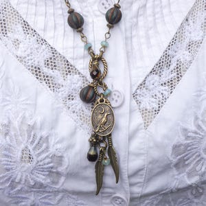 Puede incluir: Una blusa blanca de encaje con un collar largo y de capas con cuentas marrones y azules, un dije de pájaro y un colgante de metal.