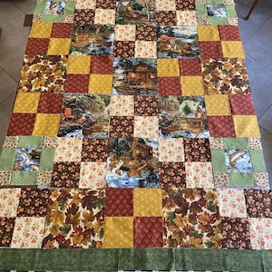 May include: A quilt top with a patchwork design featuring various scenes of nature, including cabins, trees, and fish. The quilt is made up of squares of different fabrics in shades of brown, green, yellow, and red. The quilt is laid out on a checkered table cloth.