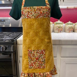 May include: A mustard yellow apron with a floral pocket and trim. The apron features a ruffle at the bottom and straps with a matching floral pattern. The apron is displayed on a mannequin in a kitchen setting.