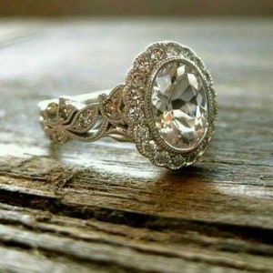 May include: A silver engagement ring with a large oval-shaped diamond surrounded by smaller diamonds. The band is intricately designed with floral motifs.
