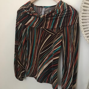 May include: A long-sleeved blouse with a cowl neck, featuring a brown background with a colorful striped pattern in red, teal, and white. The blouse is hanging on a wooden hanger against a white wall.