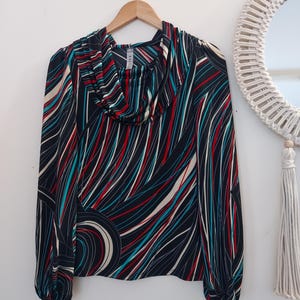 May include: A long-sleeved blouse with a cowl neck, hanging on a wooden hanger. The blouse features a black background with a swirling pattern of red, teal, and white lines. The sleeves are gathered at the wrists.