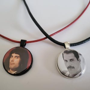 May include: Two round pendant necklaces featuring portraits of Freddie Mercury. One pendant has a black frame on a red cord with a colour portrait, while the other has a silver frame on a black cord with a black and white portrait of the musician.