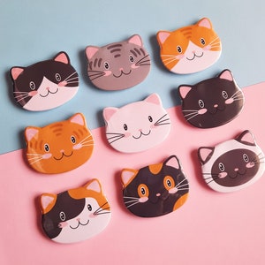 Puede incluir: Nueve imanes con forma de gato de colores con diferentes patrones y colores. Los imanes tienen un diámetro de aproximadamente 5 cm y un fondo blanco con contornos negros.