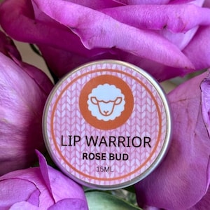 May include: A round, silver lip balm tin with the words "LIP WARRIOR ROSE BUD" printed on it. The tin has a pink background with a white sheep graphic inside an orange circle. The tin is surrounded by purple rose petals.