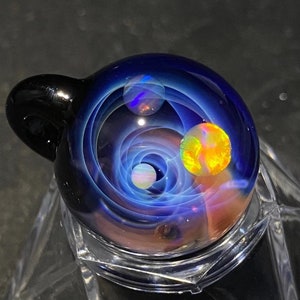 May include: A glass pendant with a swirling galaxy design. The pendant features a dark blue background with white and orange orbs, and a black loop for a chain. The glass is displayed on a clear stand.