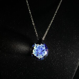 May include: A silver chain necklace with a round pendant. The pendant features a blue and white floral design encased in clear glass. The background is dark, highlighting the necklace's details.