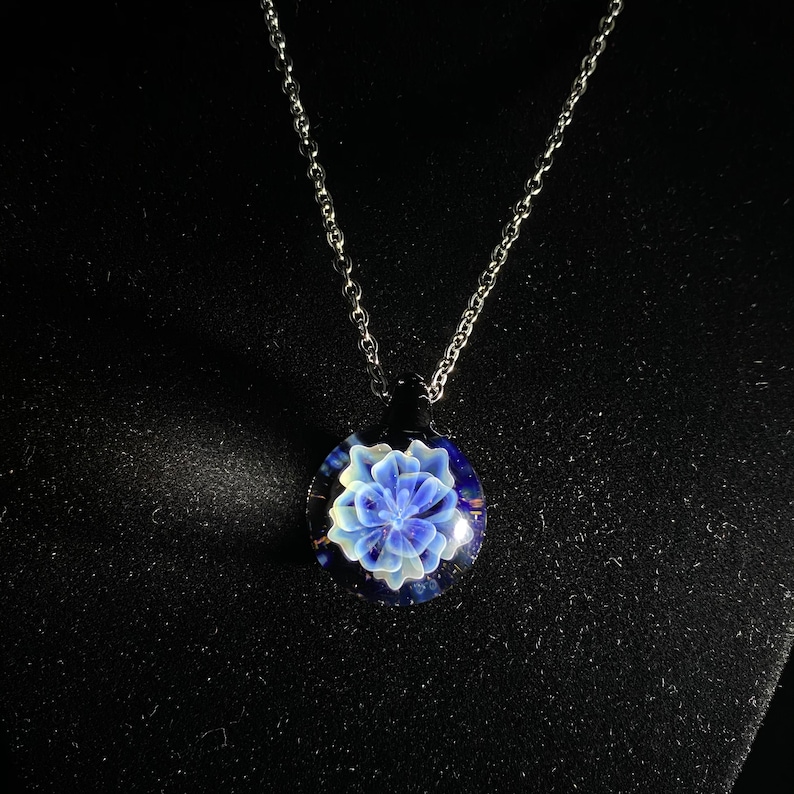 May include: A silver chain necklace with a round pendant. The pendant features a blue and white floral design encased in clear glass. The background is dark, highlighting the necklace's details.