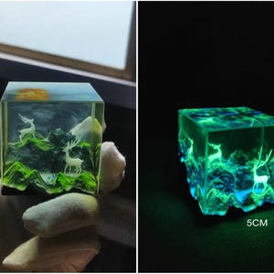 Puede incluir: Un cubo de resina transparente con una escena de paisaje en miniatura. La escena incluye ciervos blancos, colinas verdes y un sol amarillo. El cubo brilla en verde y azul en la oscuridad y mide 5 cm x 5 cm.