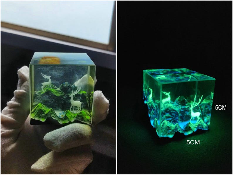 Puede incluir: Un cubo de resina transparente con una escena de paisaje en miniatura. La escena incluye ciervos blancos, colinas verdes y un sol amarillo. El cubo brilla en verde y azul en la oscuridad y mide 5 cm x 5 cm.