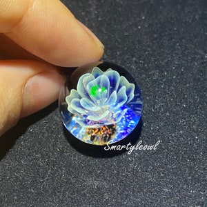 May include: A clear glass marble with a detailed, three-dimensional flower design. The flower has white and blue petals with a green center. The base of the marble has a gold and brown pattern. The text "Smartybeowl" is visible.
