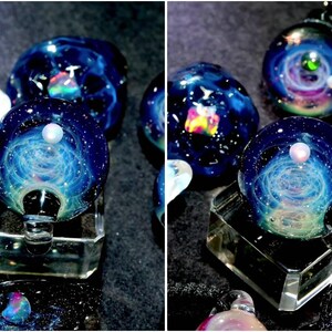 May include: Several glass pendants with a galaxy design. Each pendant is a sphere with swirling blue, white, and teal colors, resembling a nebula. Some have a small, white pearl-like bead on top. The pendants are displayed on clear, geometric stands.