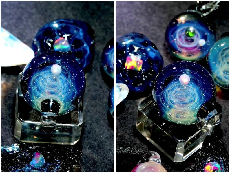 May include: Several glass pendants with a galaxy design. Each pendant is a sphere with swirling blue, white, and teal colors, resembling a nebula. Some have a small, white pearl-like bead on top. The pendants are displayed on clear, geometric stands.