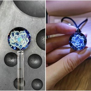 May include: A handcrafted glass pendant with a blue floral design. The pendant is round, with a dark blue background and a detailed, light blue flower at the center. The pendant is on a black cord.