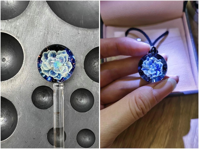 May include: A handcrafted glass pendant with a blue floral design. The pendant is round, with a dark blue background and a detailed, light blue flower at the center. The pendant is on a black cord.
