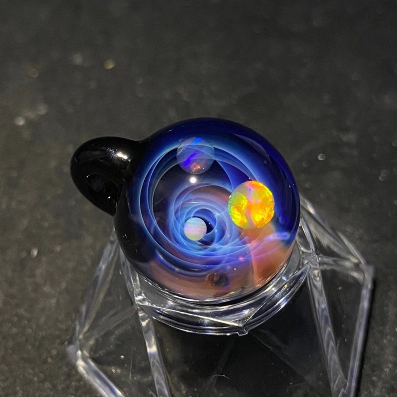 May include: A glass pendant with a swirling galaxy design. The pendant features a dark blue background with white and orange orbs, and a black loop for a chain. The glass is displayed on a clear stand.