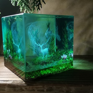 Puede incluir: Un cubo de resina transparente que muestra una escena de bosque en miniatura. Dos ciervos blancos se alzan entre plantas y flores verdes, con un efecto brumoso y et&eacute;reo. La obra de arte est&aacute; iluminada con un suave brillo verde.